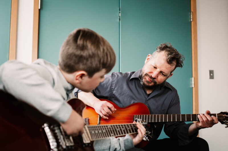 Male teacher demonstrating to male student, both holding guitars